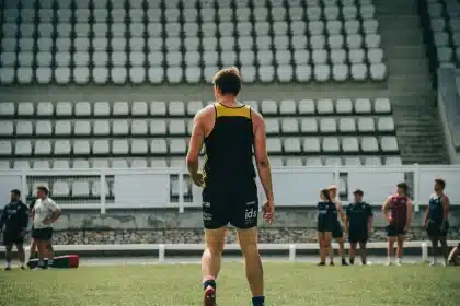 Treinador em pé no banco de suplentes durante um jogo de futebol, com estádio ao fundo.