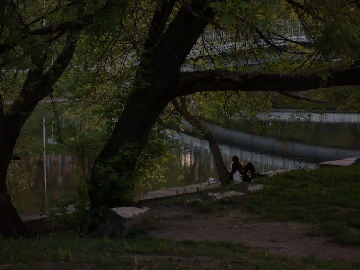 Figura solitária sentada num banco junto à margem ao entardecer, observando o rio