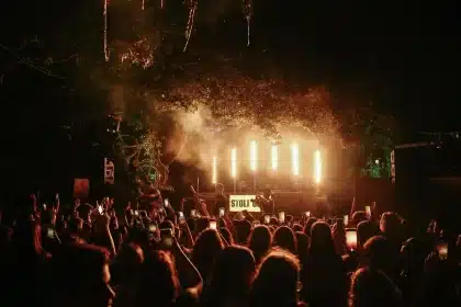 Multidão de festivaleiros a dançar e celebrar sob luzes de palco numa praia fluvial à noite