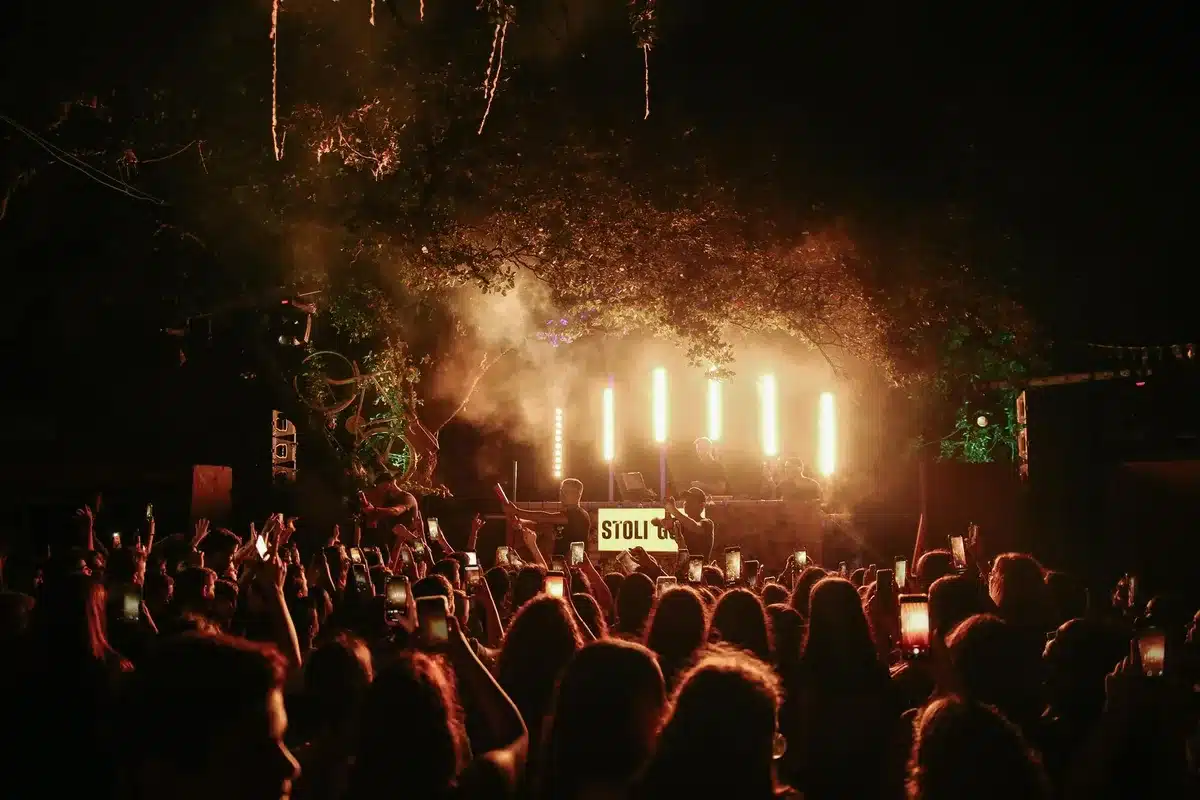 Multidão de festivaleiros a dançar e celebrar sob luzes de palco numa praia fluvial à noite