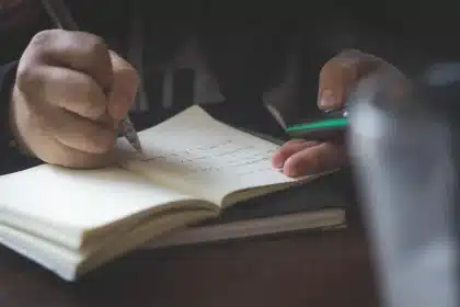 Mesa de sala de aula com caderno aberto, tablet virado para baixo e mão com lápis